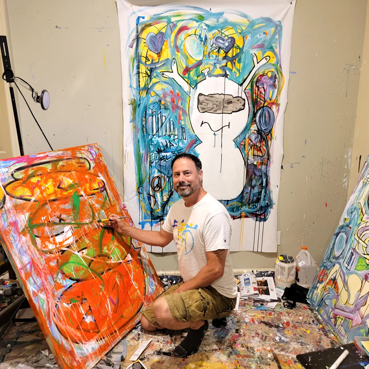 Jason Piken kneeling in his studio, painting a large orange canvas with finished work on the wall behind him